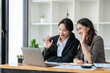 © crizzystudio - Image of two beautiful Asian businesswomen waving greeting online through a laptop at the table with a calculator, graph, and chart showing business growth data at the office.