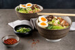 Two bowls of ramen soup with chicken breast, vegetables, mushrooms and egg on a gray concrete background and chopsticks. Front top view, healthy eating concept