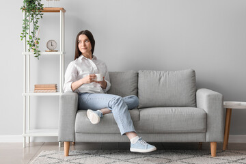 Poster - Young woman with cup of hot beverage sitting on sofa at home