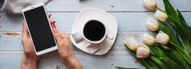 Wall Mural - bouquet of white tulips with a Cup of coffee and a smartphone in women's hand and an empty notebook on a wooden table. Concept of a spring morning workplace freelancer. Flat lay, top view composition