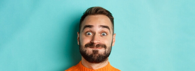 Headshot of funny bearded man showing faces and pouting, holding breath, standing in orange sweater against turquoise background