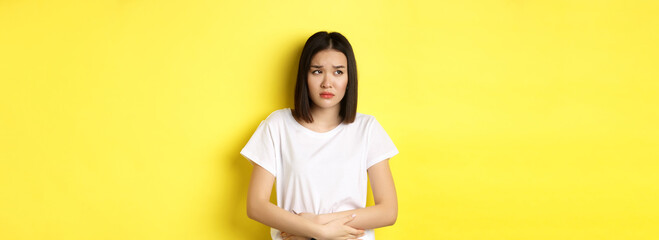 Wall Mural - Image of asian woman feeling sick, having painful cramps, holding hands on belly and frowning from pain, discomfort during menstrual period, standing over yellow background