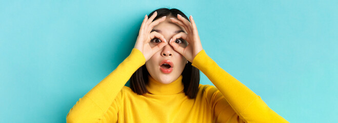 Wall Mural - Close up of funny asian woman looking through hand binoculars with surprised face, see something amazing, standing over blue background