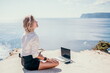 © panophotograph - Woman sea laptop yoga. Business woman freelancer in yoga pose working over blue sea beach at laptop and meditates. Girl relieves stress from work. Freelance, digital nomad, travel and holidays concept