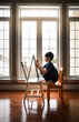 © Cavan Images - Young boy drawing on a canvas on an easel in front of windows.