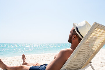 Wall Mural - Young man in white hat relaxing in deck chair on beautiful sandy beach.