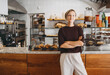© nataliaderiabina - Portrait of smiling young woman entrepreneur standing at the counter of her bakery and coffee shop. Local small business owner indoors.