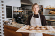 © nataliaderiabina - Welcoming female baker holding freshly baked almond croissants in  background of bakery shop.
