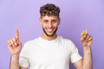 Wall Mural - Young caucasian man holding a Bitcoin isolated on purple background showing and lifting a finger in sign of the best