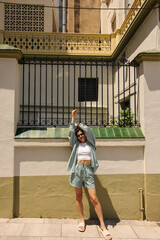 Wall Mural - In full length positive young caucasian woman in sunglasses stretches hands up, looking at camera. Brunette wears white singlet, green suit and slippers. Concept leisure vacation.