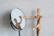 A Mannequin Wooden Man Stands On A Light Background And Looks In The Mirror.