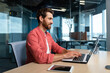 © Liubomir - Successful businessman in red shirt happily working with laptop inside office, mature man with beard at workplace typing on keyboard smiling satisfied with work results and achievement.