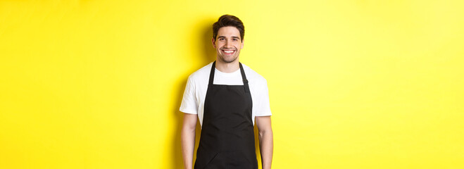 Wall Mural - Young man waiter in black apron, smiling, working in store or coffee shop, standing against yellow background