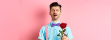 Heartbroken Guy In Funny Bow-tie Crying Over Girlfriend, Standing Alone With Red Rose On Pink Background And Sobbing, Break-up On Valentines Day