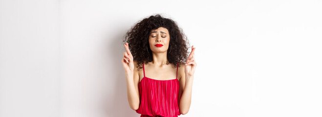 Hopeful woman in red dress making wish, praying with fingers crossed and closed eyes, pleading god, standing over white background