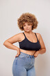 © South House Studio - Confident plus size woman with Afro hair feels confident, wears jeans and black lingerie bra, poses in studio over white wall