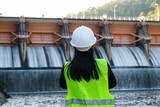 Fototapeta Uliczki - Rear view of female engineer in green vest and helmet standing outside against background of dam with hydroelectric power plant and irrigation.