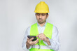© anut21ng Stock - Asian male construction worker in reflective vest and yellow safety helmet holding smartphone, utilizing internet social media in construction industry