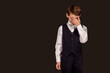 Little boy in suit with hand covering face standing on black isolated background, wear white shirt and bow tie. Schoolboy student posing in studio. Education concept. Copy text space for advertising