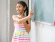 © Nassorn - Smart girl student pointing at a blackboard in an elementary school classroom. Portrait of schoolgirl standing at chalkboard smiling. Cheerful child back to school for cognition education concept.