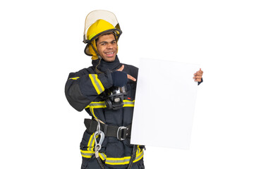 Wall Mural - Young smiling African American fireman wearing uniform and helmet looking to the camera and holding blank paper sheet in hand and finger pointing. Copy space for text, isolated on white background 