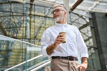 Confident senior mature old successful business man leader, smiling mid aged senior professional businessman executive ceo wearing white shirt holding coffee looking away thinking standing outdoor.