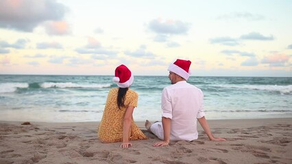 Wall Mural - Young couple in red Santa hats sitting on the beach on Christmas