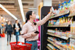 © _KUBE_ - Side view of Caucasian woman choosing products in grocery store. Student reaches hand to top shelf holds cellphone and cart. Shelves with food in background. Concept of shopping and consumerism