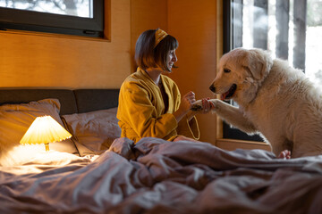 Wall Mural - Young woman cares her huge adorable white dog while lying in bed in tiny bedroom of wooden cabin on nature. House coziness and friendship with pets concept