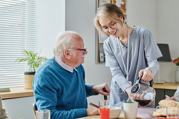 Young caregiver pouring coffee from teapot for senior man while he having breakfast at table