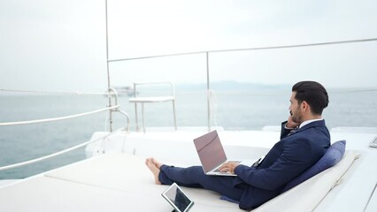 Wall Mural - Businessman working on laptop computer and talking on mobile phone for global corporate business while travel on luxury private catamaran boat yacht sailing in the ocean on summer vacation at sunset.