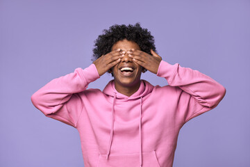 Wall Mural - Happy African American teen guy closing eyes with hands feeling amazed waiting for surprise. Smiling blind ethnic student model excited with anticipation standing isolated on light purple background.