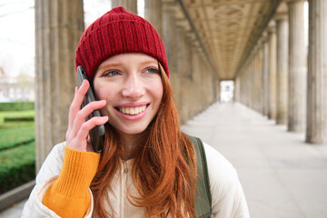 Portrait of redhead european girl in red hat, makes a phone call, walks in city and talks to friend on smartphone