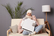 © sementsova321 - Indoor shot of smiling happy positive woman wearing cap and winter coat sitting on the sofa at home and embracing heater, looking at camera, waiting for turning on central heating.