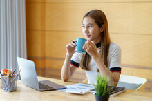 Pretty Asian Businesswoman Sitting Holding Coffee At Home Working Smiling And Enjoying Working With Laptop Happily.