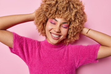 Wall Mural - Happy curly haired young European woman wears vivid pink makeup smiles toothily keeps eyes closed wears t shirt keeps hands behind head expresses positive emotions isolated over rosy background.