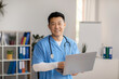 © Prostock-studio - Cheerful middle aged korean doctor in uniform with stethoscope with computer in clinic office interior