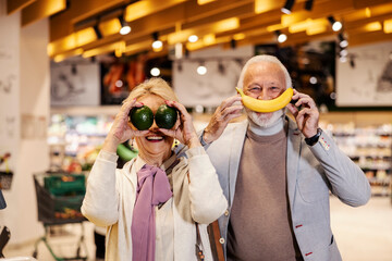 Wall Mural - A silly senior couple is pretending fruits are their mouth and eyes while standing at the supermarket.