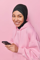 Wall Mural - Happy islamic woman dressed in casual hoodie and black hijab holds mobile phone uses application for chatting online looks with joy at camera isolated over pink background. Technology and religion