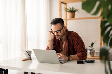 Focused young businessman using laptop working online in a home office. Professional businessman looking at laptop checking web market, e-learning webinar, having remote hybrid call sitting indoors.