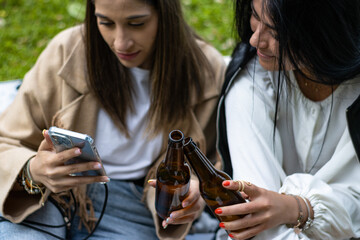 Two smiling female friends toasting with beer sitting on the grass. Lifestyle of young female friends sitting on the grass toasting with beer bottles