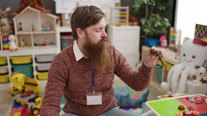 Young redhead man preschool teacher stressed sitting on chair at kindergarten