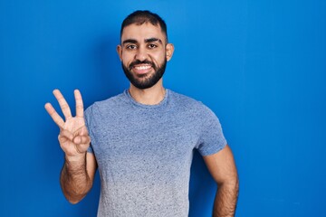 Wall Mural - Middle east man with beard standing over blue background showing and pointing up with fingers number three while smiling confident and happy.