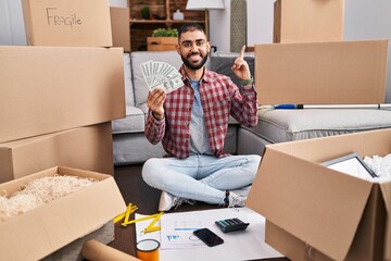 Sticker - Middle east man with beard sitting on the floor at new home holding money surprised with an idea or question pointing finger with happy face, number one