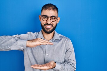 Wall Mural - Middle east man with beard standing over blue background gesturing with hands showing big and large size sign, measure symbol. smiling looking at the camera. measuring concept.