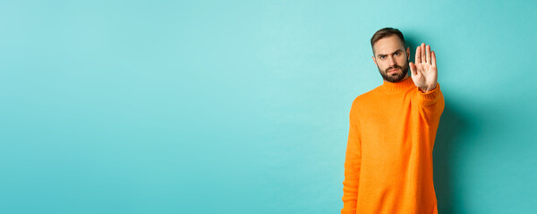 Serious frowning man showing stop gesture, telling no, prohibit and disagree, standing over light blue background