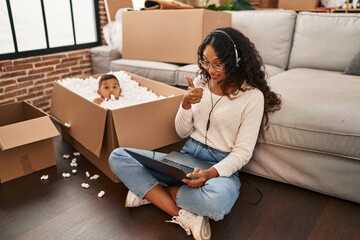 Poster - Young hispanic mother and kid sitting on the floor at new home using laptop smiling happy and positive, thumb up doing excellent and approval sign