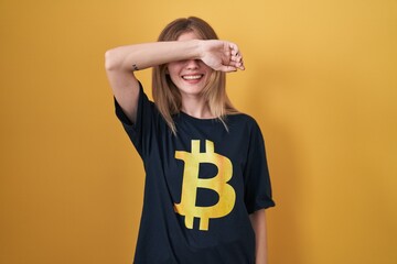 Poster - Blonde caucasian woman wearing bitcoin t shirt smiling cheerful playing peek a boo with hands showing face. surprised and exited