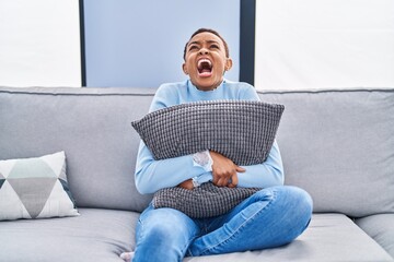 Sticker - African american woman sitting on the sofa at home hugging pillow angry and mad screaming frustrated and furious, shouting with anger looking up.