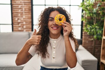 Young hispanic woman holding tasty colorful doughnut  eye smiling happy and positive, thumb up doing excellent and approval sign